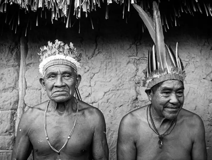 Dois anciãos com cocar, diante de uma casa de barro.